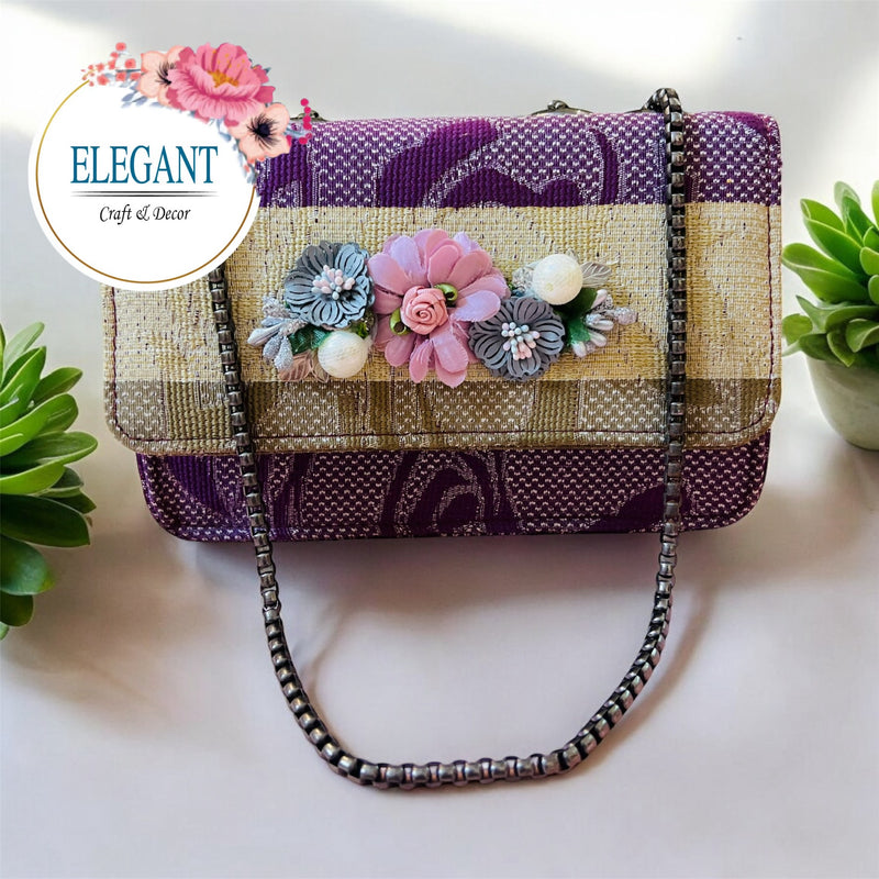 A vibrant, gold-and-purple patterned envelope clutch adorned with a centered row of detailed 3D flowers and a metallic chain strap.