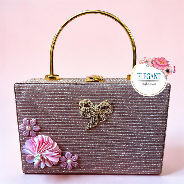 Elegant, rectangular box-style clutch featuring a shimmering, textured fabric in a mauve or rose-gold tone. It is designed with a striking gold-toned circular handle and a secure top clasp.